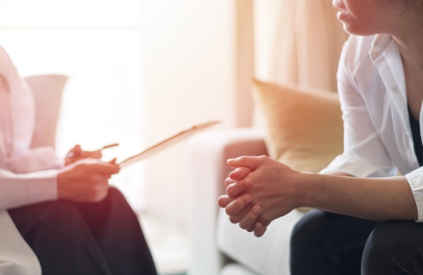 A person sitting on a couch with their hands clasped, facing their therapist. Their therapist is holding a clipboard.